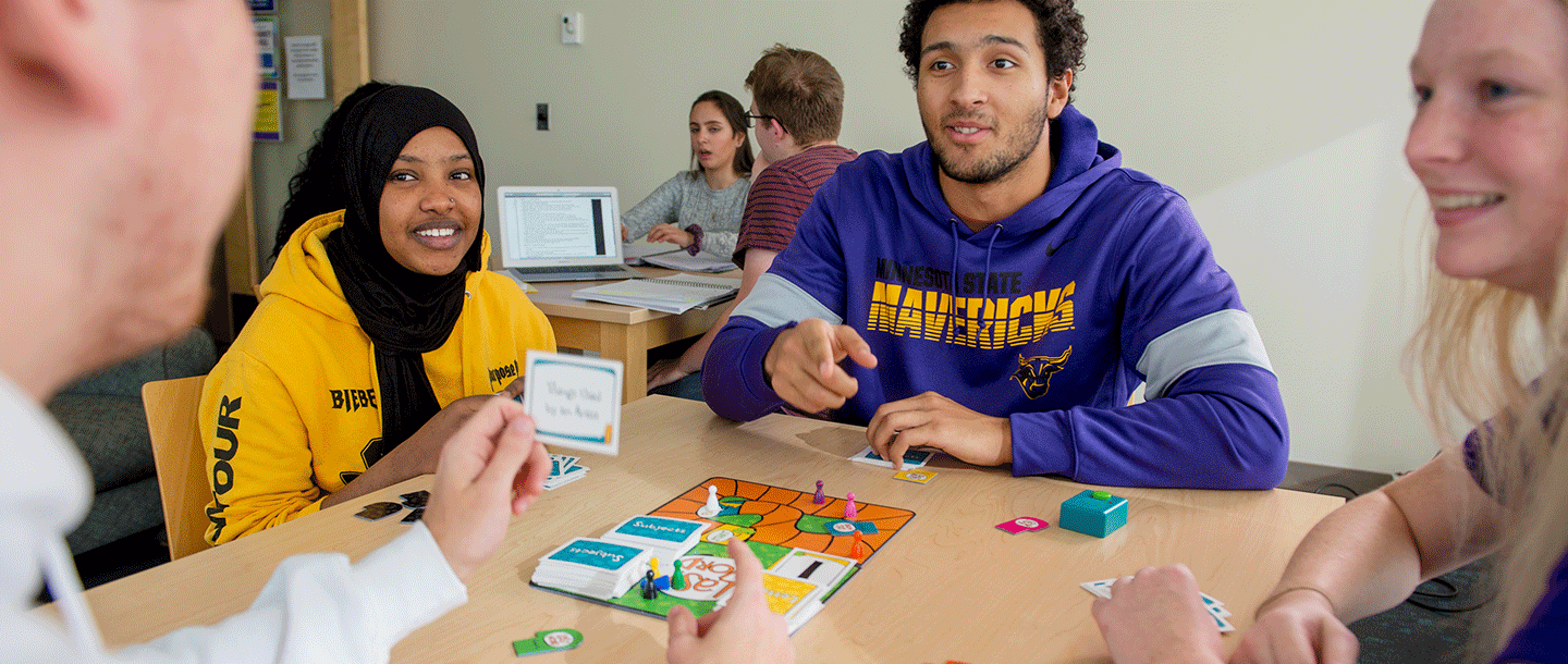 Four students sitting at a table in a room playing a board game while two students are sitting at another table behind them studying with a laptop